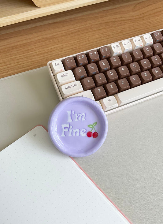 This cute tray dish has so many fun purposes. You can use this to hold your jewelry, simply use as a decor piece in your room or on your desk, or even as a mini coaster for a small can. This design is a light purple base, features the verbiage "I'm Fine" and has cherries painted on.